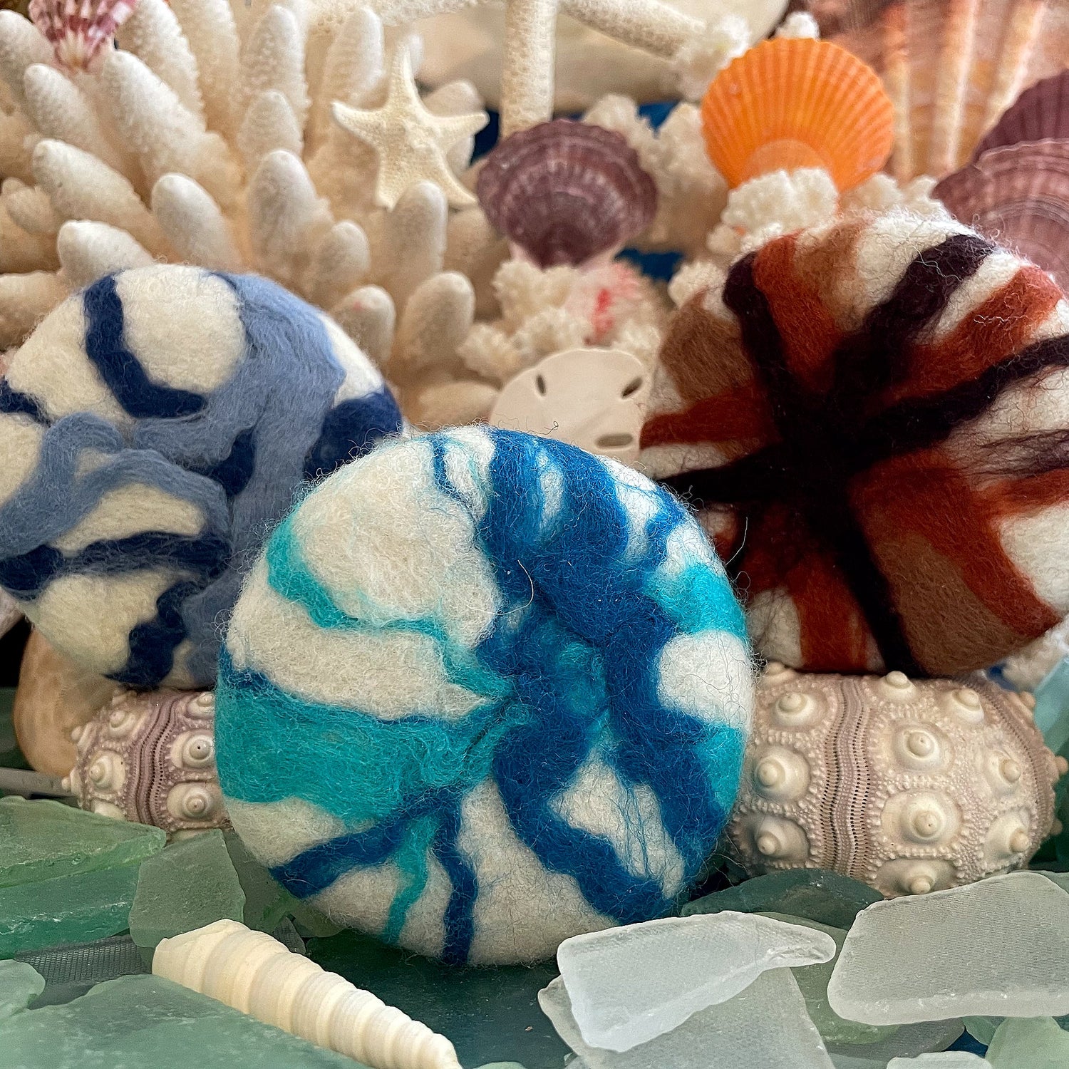 Assortment of blue, brown, and white felted olive oil soaps resting on sea glass and coral, representing the variety of Whale Tale Soapworks’ artisan collection.