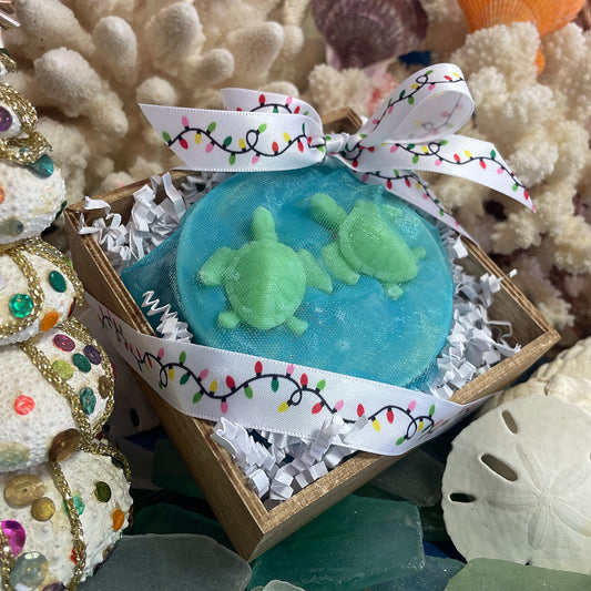 Turtle Tracks soap gift box featuring a round ocean-blue soap with two small turtle-shaped embeds, wrapped in mesh and tied with a holiday light ribbon.